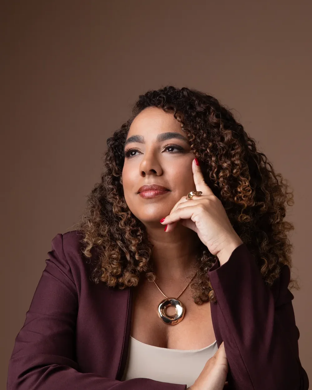 Retrato de mulher de blazer vinho, cabelos cacheados e mão próxima ao rosto