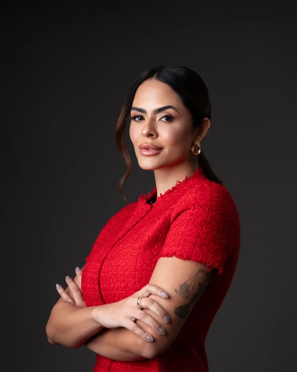 Retrato de mulher com roupa vermelha e braços cruzados diante de fundo escuro