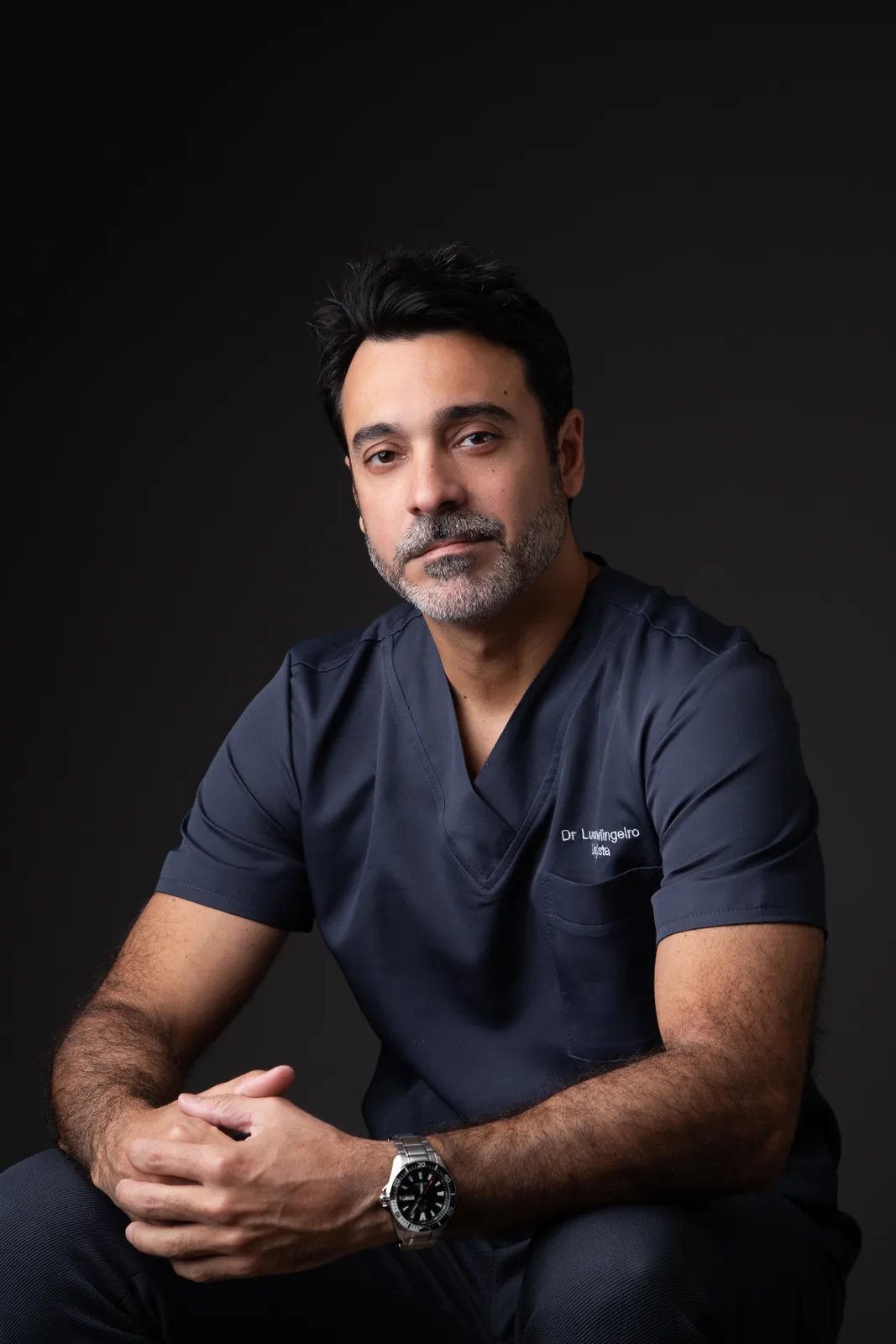 Retrato profissional de médico sentado com roupa cirúrgica azul-marinho