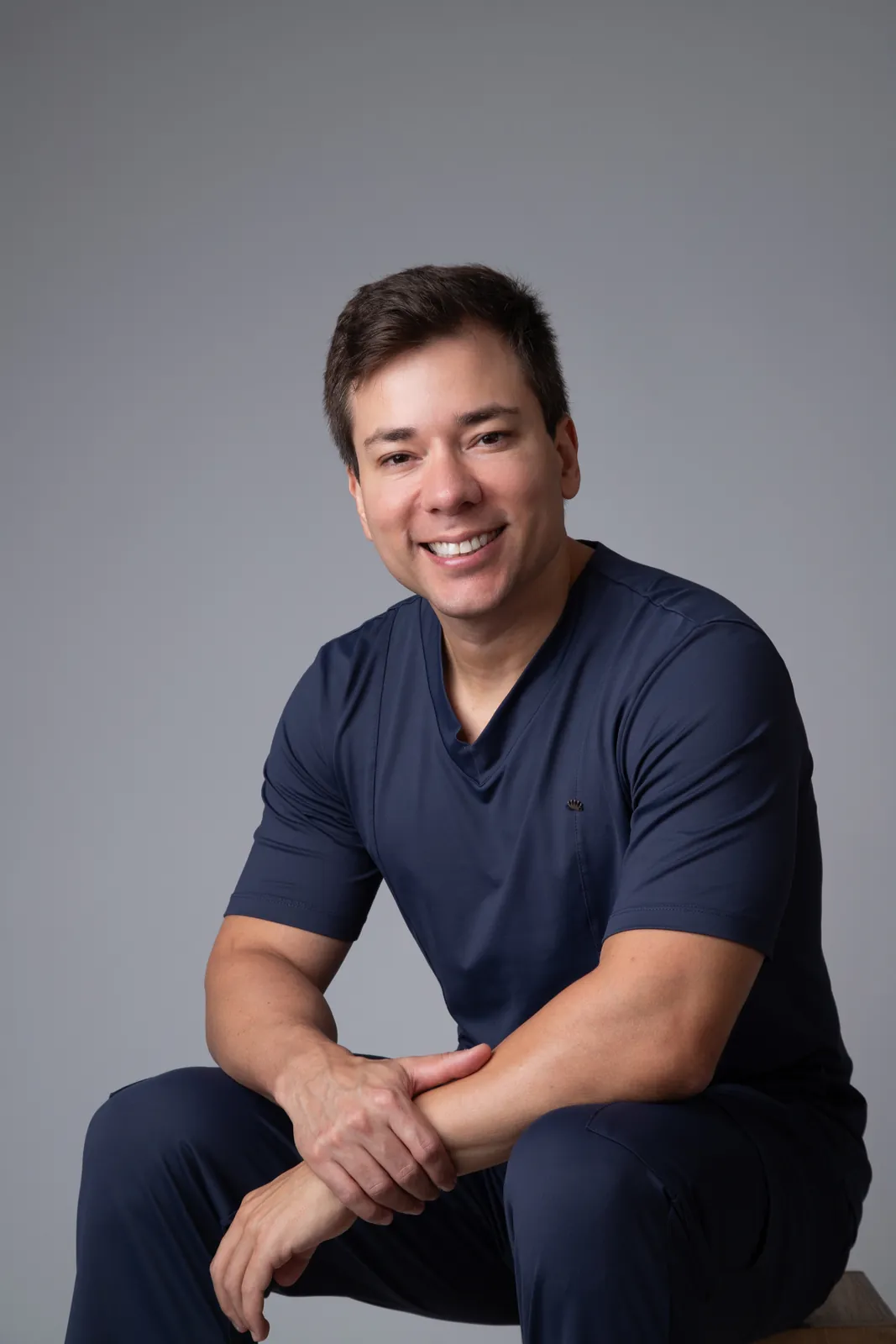 Retrato profissional de médico sorrindo com roupa cirúrgica azul-marinho sentado em banqueta