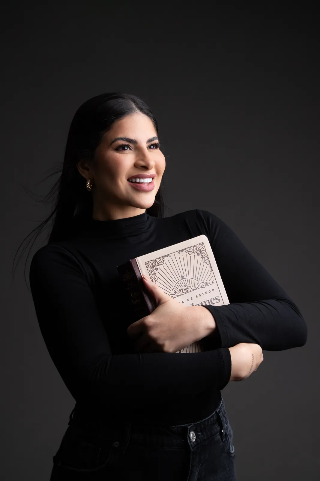 Retrato de autora sorrindo com blusa preta segurando um livro sobre fundo escuro